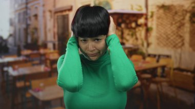 Restoran terasında kulaklarını kapatan yeşil tişörtlü kadın arka planda oturan stresli ya da bunalmış görünüyor..