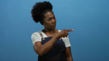 Önlük takan kadın mavi arka planda özgüvenli bir şekilde odak ve yönünü izole bir ortamda somutlaştırıyor, iddialı hareketlerle fikirleri işaret ediyor..