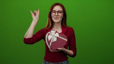 Taş gibi hareketlerle gülümseyen bir kadın elinde kalp şeklinde bir hediye kutusuyla gençlik tarzını sergileyen yeşil arka planda..