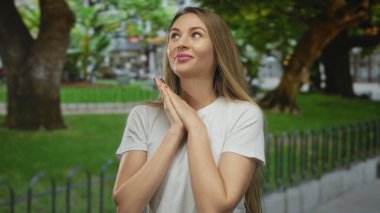 Yeşil bir parkta, arka planda ağaçlar olan, elleri bağlı gülümseyen bir kadın, açık havada güzel bir günün tadını çıkarıyor..