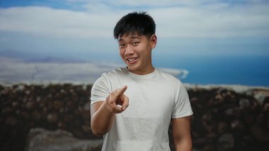 Genç Çinli bir adam arka planda okyanus manzaralı deniz kenarında neşeli bir jest yapıyor, beyaz bir gömlek giyiyor ve açık yürekli bir tavır sergiliyor.