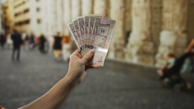 Suudi Arabistan riyal paralarını şehir sokaklarında elinde tutan adam, şehir içi dolandırıcılık, seyahat ve finans kavramlarını canlı bir şekilde gösteren para biriminin detaylarını sergiliyor..