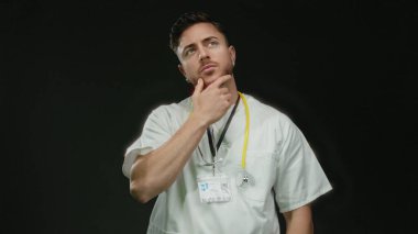 Sakallı ve steteskoplu genç bir adam siyah bir arka plana karşı düşünceli bir şekilde duruyor. Tecrit edilmiş modern bir tıp uzmanını somutlaştırıyor..