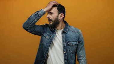 Kot gömlekli İspanyol sakallı adam canlı turuncu arka planda düşünceli bir ifade ve enerjik bir hareket sergiliyor..