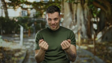 İspanyol bir adam parkta bir jest yapıyor, dövmeleri var ve yeşil bir gömlek giyiyor, doğal ortamda cazibe ve güven yayıyor..