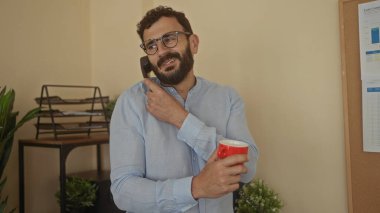 Kırmızı bardakta sakallı İspanyol bir adam ofis ortamında telefonla konuşuyor. Etrafı bitkiler ve evraklarla çevrili. Nişanlı ve profesyonel görünüyor..