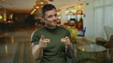 Genç adam gösterişli otel lobisinde gülümsüyor sanatsal dövmeleri ve şık ışıklandırma ve dekorasyonun ortasında günlük kıyafetleriyle özgüven ve cazibesini iletiyor..