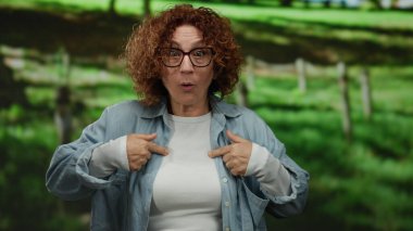 Gözlüklü bir kadın yeşil bir parkta dikilirken şaşkınlıkla kendini işaret ediyor..