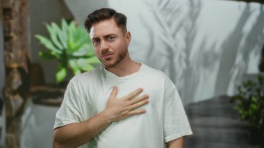 Şehir sokağında öksüren genç bir adam, beyaz bir gömlek giyiyor ve yeşil bir ortamda göğsünde bir elle endişe gösteriyor..