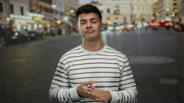 Genç adam hareketli şehir caddesinde sıradan çizgili gömleğiyle, canlı şehir gece lambalarıyla çevrili duruyor.