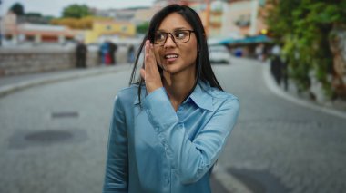 Mavi gömlekli genç bir kadın canlı bir şehir caddesinde fısıldıyor, renkli binaların arka planına sahip samimi, neşeli bir açık hava anını yakalıyor..