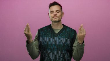 Genç İspanyol bir adam, yeşil bir süveter giyerken sakin ve odaklanmış bir şekilde, izole edilmiş pembe bir arka plan önünde meditasyon yapıyor.