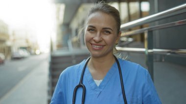 Gülümseyen hemşire üniformalı kadın şehir sokağında büyüleyici bir gülümseme ve sarı saçlarıyla kliniğin önünde profesyonel ve misafirperver bir tavır sergiliyor..