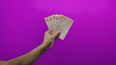 Türk lira banknotları tutan adam, canlı mor bir arka plana karşı, ekonomik ve finansal etkileri ile elde para sergileniyor.