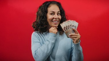 Meksika pesolarını elinde tutan kadın canlı kırmızı bir arkaplan karşısında gülümsüyor, zenginliği ve finansal temayı sembolize ediyor.