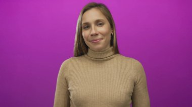 Canlı pembe bir arka plana gülümseyen, kahverengi bir kazak giyen, sakin ve kendinden emin bir ifade sergileyen bir kadın..