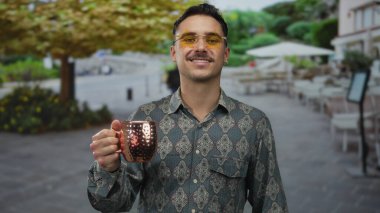 Genç adam gülümsüyor ve canlı bir şehir caddesinde fincanı tutuyor, şık gözlükler takıyor ve gür ağaçların altında desenli gömlek giyiyor, rahat bir şehir havası yayıyor..