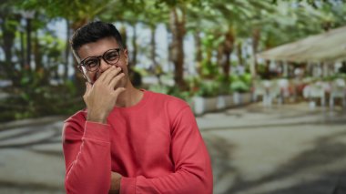 Güneşli bir terasta, hareketli bir açık hava restoranında gülümseyen genç bir adam, canlı bir şehirde mutluluk ve rahatlama yayıyor..