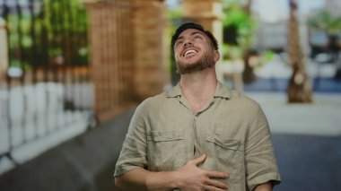 Sakallı genç adam, günlük tişörtler giymiş, şehir dışında arka planda yakalanmış, mutluluk ve rahatlama yayıyor..