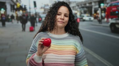 Şehir caddesinde kalp şeklinde bir nesne tutan kadın renkli bir kazak giyiyor, canlı bir ifade ve şehir hayatı yansıması yakalıyor..