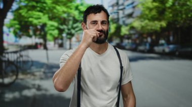 Sakallı İspanyol bir adam gün ışığında şehir dışındaki bir sokakta gizli bir jest yapıyor. Etrafı bulanık yeşil ağaçlar ve binalarla çevrili..