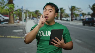 Yeşil tişörtlü genç adam şehir merkezinde bulutlu gökyüzüne karşı tablet tutarken düşünceli bir ifade ve günlük tarzın vücut bulmuş hali.