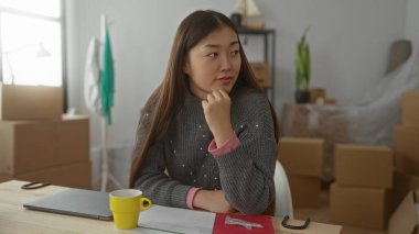 Asyalı bir kadın yeni bir evde oturuyor. Etrafı kutularla çevrili, gülümseyen ve düşünceli bir şekilde bir masada dizüstü bilgisayar ve kahve fincanıyla oturuyor..