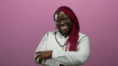 Saçları örgülü, gözlüklü, kolları çapraz pembe bir arkaplan ile gülümseyen, beyaz gömlek giyen, mutluluk ve pozitifliği ifade eden kendinden emin Afro-Amerikan kadını..
