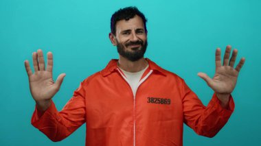 Turuncu hapishane tulumu giyen İspanyol sakallı adam mavi arka planda duruyor. Elleri havada ve sakin bir şekilde teslim ol ya da dur senaryosu öneriyor..