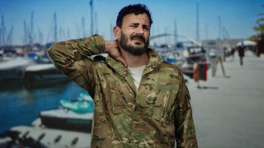 İspanyol üniformalı, sakallı bir adam tekneleri görünen hareketli bir limanda duruyor, parlak mavi gökyüzünün altında boynuna masaj yapıyor gibi görünüyor..