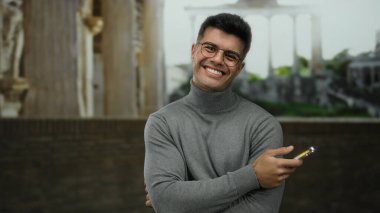 Arkaplanda Roma harabeleri olan akıllı telefona gülümseyen genç bir adam tarihi açık hava ortamında modern teknoloji kontrastı sergiliyor..