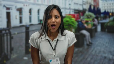 Kalabalık bir caddede, dışarıda duran genç bir kadın. Şaşırmış bir ifadeyle, beyaz bir gömlek ve kimlik kartı takıyor. Şehirde çeşitli duygular sergiliyor..