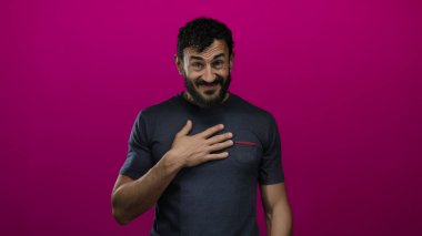 Sakallı İspanyol bir adam canlı pembe arka plana karşı şaşkınlık ve minnettarlık ifade ediyor, elleri göğsünde, duyguyu çağrıştırıyor, günlük koyu tişört giyiyor..