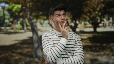 Çizgili tişörtlü, düşünceli bir genç adam güneşli bir parkta etrafı ağaçlarla çevrili, sakin bir açık hava manzarası sergiliyor..