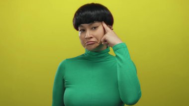 Yeşil kazaklı Çinli kadın, sarı arka planda düşünceli bir şekilde poz veriyor. Zekâsını şakakta sergiliyor..