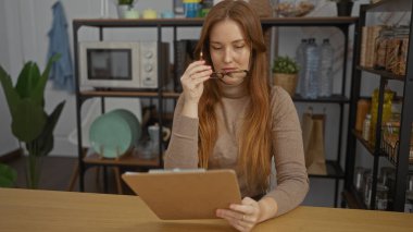 Ofiste gazete kupürleri okuyan kadın, kızıl saçlı, günlük kıyafetler, modern iç mekan ve odaklı ifade, organize raf ve çevre mobilyaları ile çevrili..