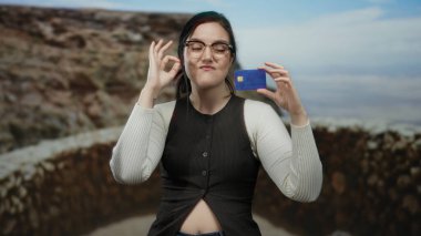 Bir kadın, kumsalda elinde kredi kartı tutarken güzel bir deniz manzarası karşısında kendine güvenini ve tarzını sergiliyor. Modern finansal özgürlüğü resmediyor..