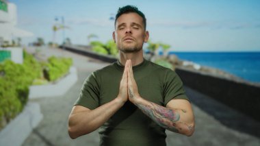 Zeytin rengi tişörtlü genç adam, deniz kenarında sakin sahiller ve okyanus arkaplanlarıyla meditasyon yapıyor, berrak gökyüzü altında huzurlu bir ifade ve sakinlik sergiliyor.