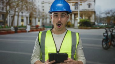 Yansıtıcı yelek ve şapka giyen genç İspanyol adam, şehir caddesinde tablete şaşırmış görünüyor, inşaat temasını ve şehir merkezini gözler önüne seriyor.
