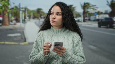 Sokakta akıllı telefon kullanan bir kadın kıvırcık saçlı bir kazak giyiyor. Odaklanmış ifade, gündüz vakti şehir ortamında teknolojiyi gösteriyor.