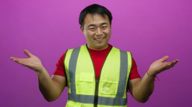 Emniyet yeleği giyen genç adam avuçlarını pembe arka plana dayamış gülümsüyor.