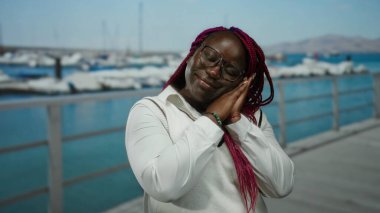 Rıhtımda gözleri kapalı bir kadın uyku jesti taklidi yapıyor, gözlük ve örgü takıyor, arka planda kayıklar ve deniz var..