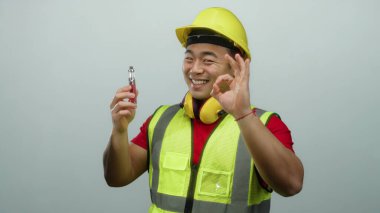 İnşaat malzemeleri ve beyaz arka planda sert bir şapka takan genç adam kendine güvenen bir gülümseme ve onaylanmış bir jest içeren bir cihaz sergiliyor.