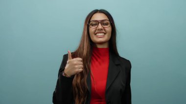 Kırmızı gömlekli ve siyah ceketli İspanyol kadın mavi izole edilmiş arka plana başparmak işareti vererek pozitifliğini ve güvenini dile getiriyor..