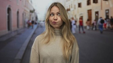 Sarışın kadın kaldırım taşı bir sokakta çenesinin altında kapalı el ele tutuşurken göz kırpar; şakacı merak neşe büyüsü.