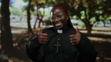 Çapraz gülümseyen ve parkın düzeninde baş parmak işareti yapan, gözlüklü ve siyah cüppeli bir kadın, doğal çevrede pozitif hava yayıyor..