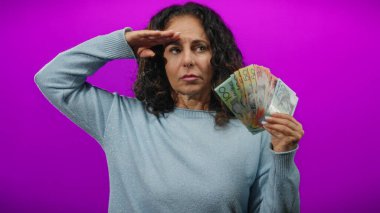 Avustralya dolarlarını canlı mor bir duvarın önünde tutan kadın, finansal güven ve orta yaş bilgeliğini göstererek selamlayıcı bir jest yapıyor..