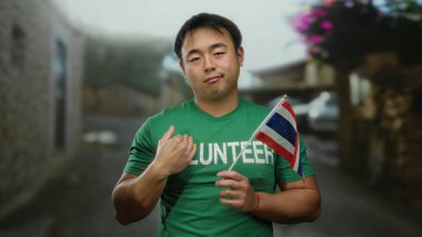 Sokaklarda Tayland bayrağı taşıyan genç adam gönüllü tişört giyiyor. Şehir ortamında kamu hizmeti öneriyor.