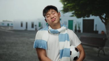 Beyaz gömlekli genç adam futbol topunu dışarıda tutarken aynı zamanda şehir caddesinde ağaca yaslanıyor ve gün boyunca sakin bir ifadeyle eşarp takıyor.