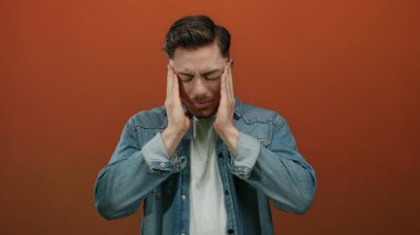 Hüsrana uğramış sakallı genç adam izole edilmiş kırmızı arka plan duvarına karşı rahatsızlık içinde başını tutuyor, baş ağrısı veya stresi ifade ediyor..